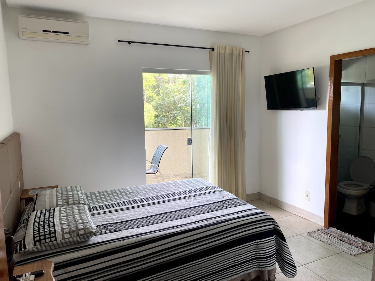 A spacious bedroom features a large bed with a striped blanket. A television is mounted on the wall opposite the bed. A glass sliding door provides access to a balcony, with natural light illuminating the room. A bathroom is visible through a partially open door.