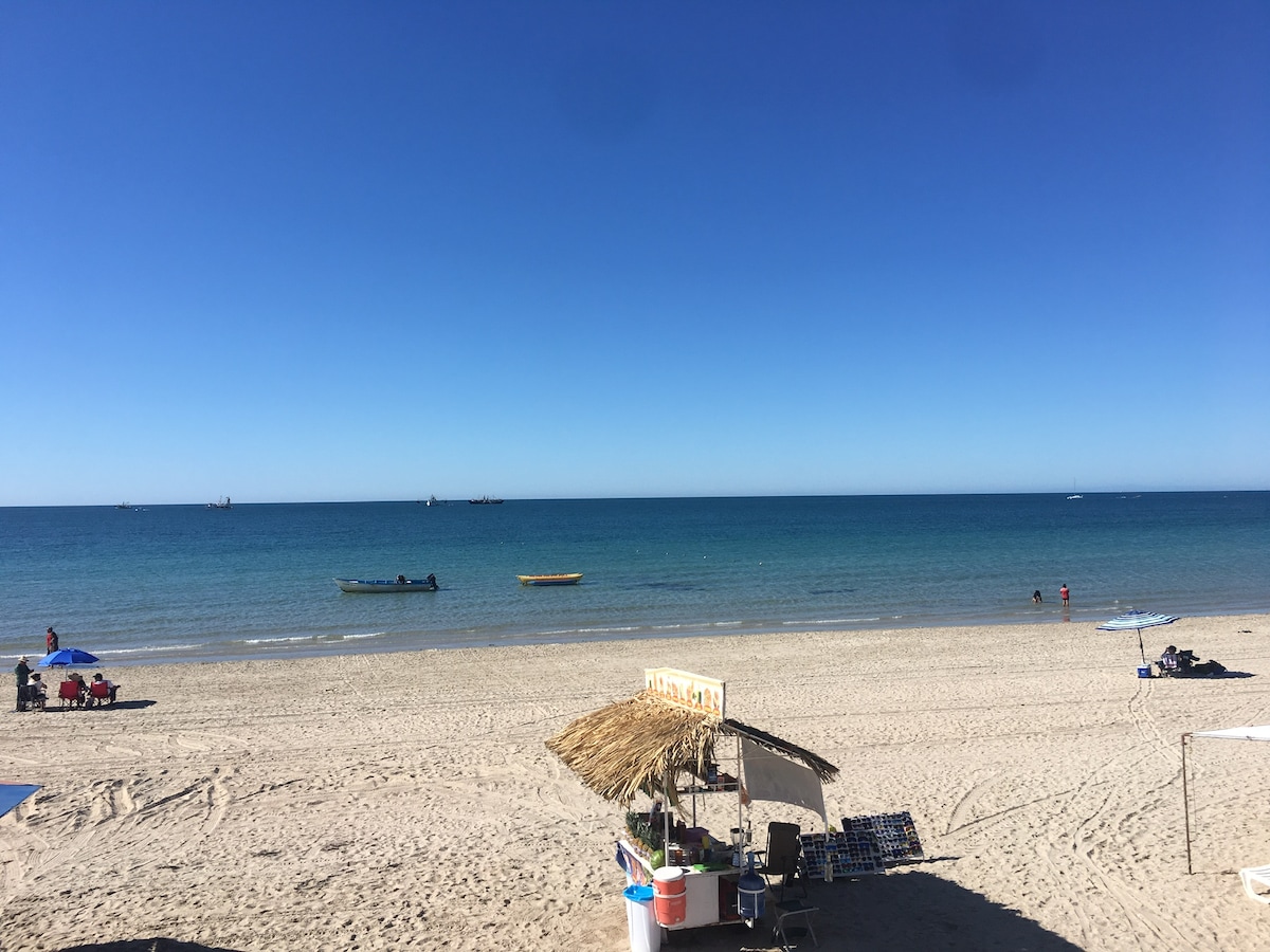 A serene beach scene features soft golden sand stretching towards calm, clear waters under a clear blue sky. Sunlight reflects off the sea, while a few boats can be seen in the distance, and beachgoers relax beneath colorful umbrellas nearby.