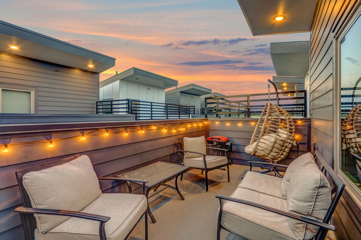 A private rooftop patio features comfortable seating with two armchairs and a coffee table, surrounded by string lights that enhance the evening ambiance. A hanging chair adds a cozy touch, while a grill provides an option for outdoor cooking.