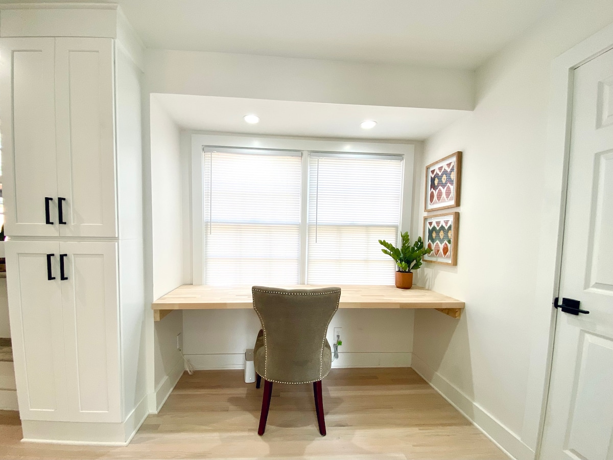 A dedicated office space is featured with a wooden desk positioned beneath bright windows, allowing ample natural light. A comfortable chair is placed at the desk, accompanied by framed decorative art and a potted green plant. Soft light fixtures enhance the workspace ambiance.