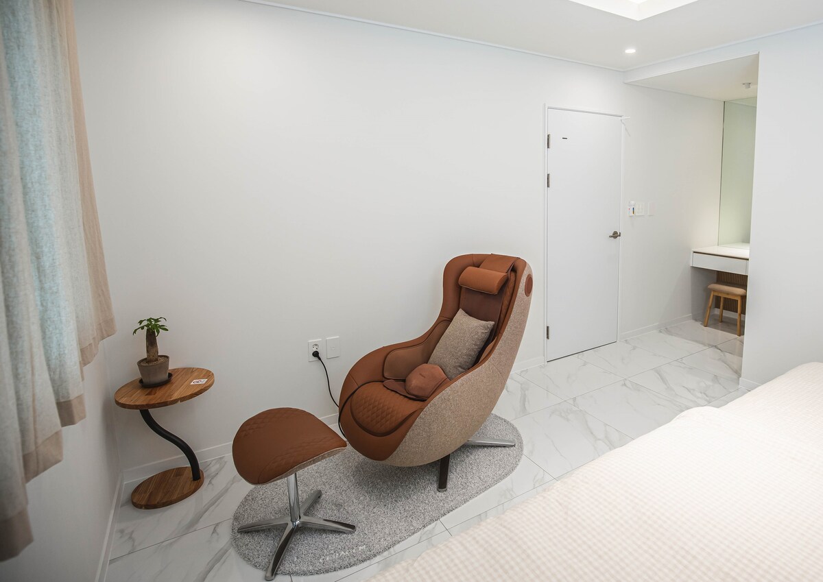A contemporary seating area features a stylish lounge chair with a matching ottoman, positioned beside a small wooden side table. A potted plant sits atop the table. The room's neutral tones and smooth tile flooring contribute to a clean and modern aesthetic.