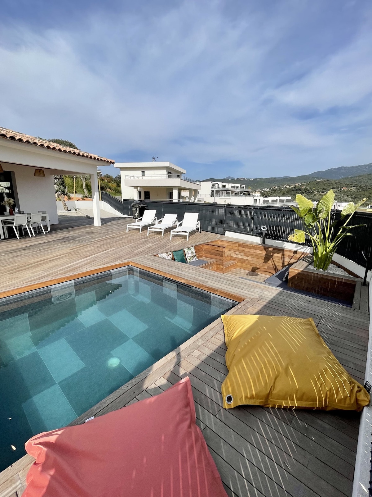 An outdoor terrace features a heated pool surrounded by wooden decking. Colorful bean bags are placed for relaxation, while poolside loungers provide additional seating. Lush greenery is visible nearby, complementing the serene atmosphere, with distant hills framed against a clear sky.