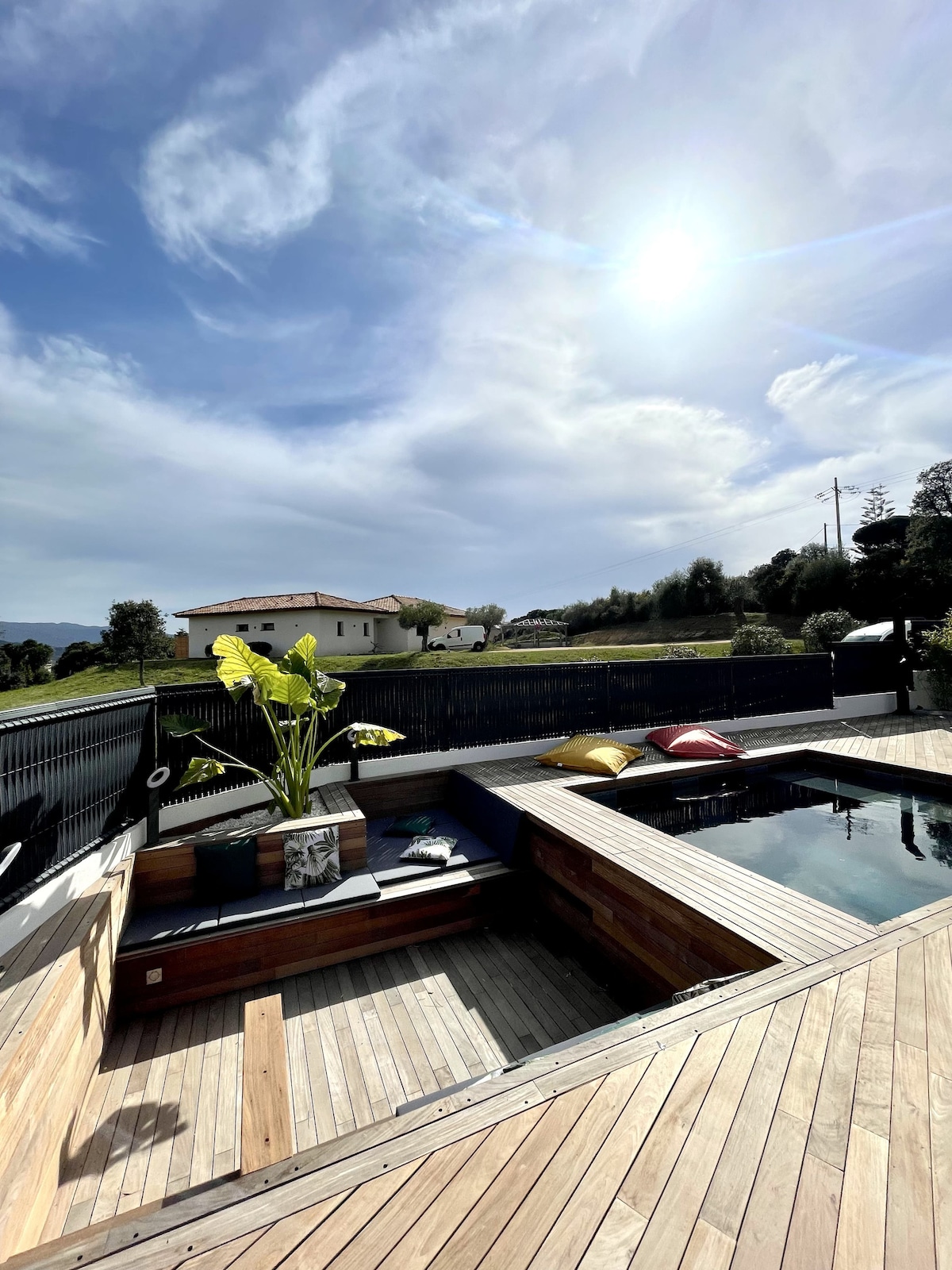 An outdoor space featuring a stylish wooden deck surrounding a heated pool. Colorful cushions are scattered around the seating area, complemented by a potted plant. The expansive sky above contains scattered clouds, with sunlight bathing the area in a warm glow.