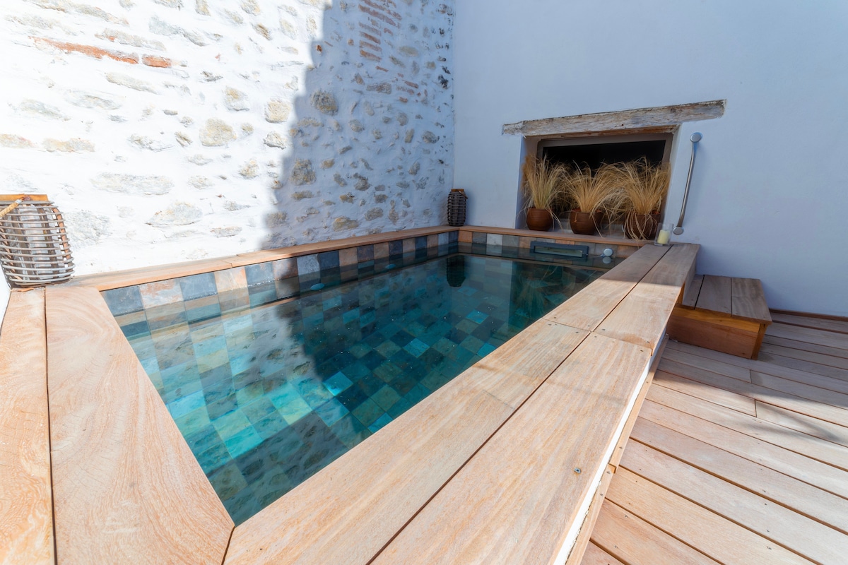 A small pool is set within a wooden deck, surrounded by a rustic stone wall. The water is clear, reflecting light and creating a tranquil space. Planters with dry grasses add a natural touch to the serene environment.