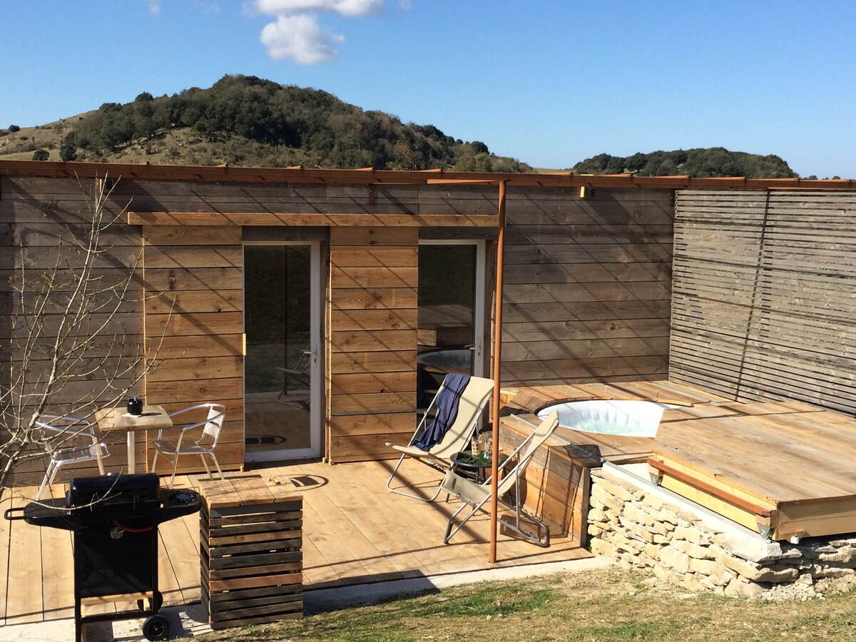 The image showcases a rustic wooden container residence nestled in a natural setting. A private jacuzzi is positioned on the wooden deck, with two chairs and a small table nearby. Mountains are visible in the background under a clear blue sky.