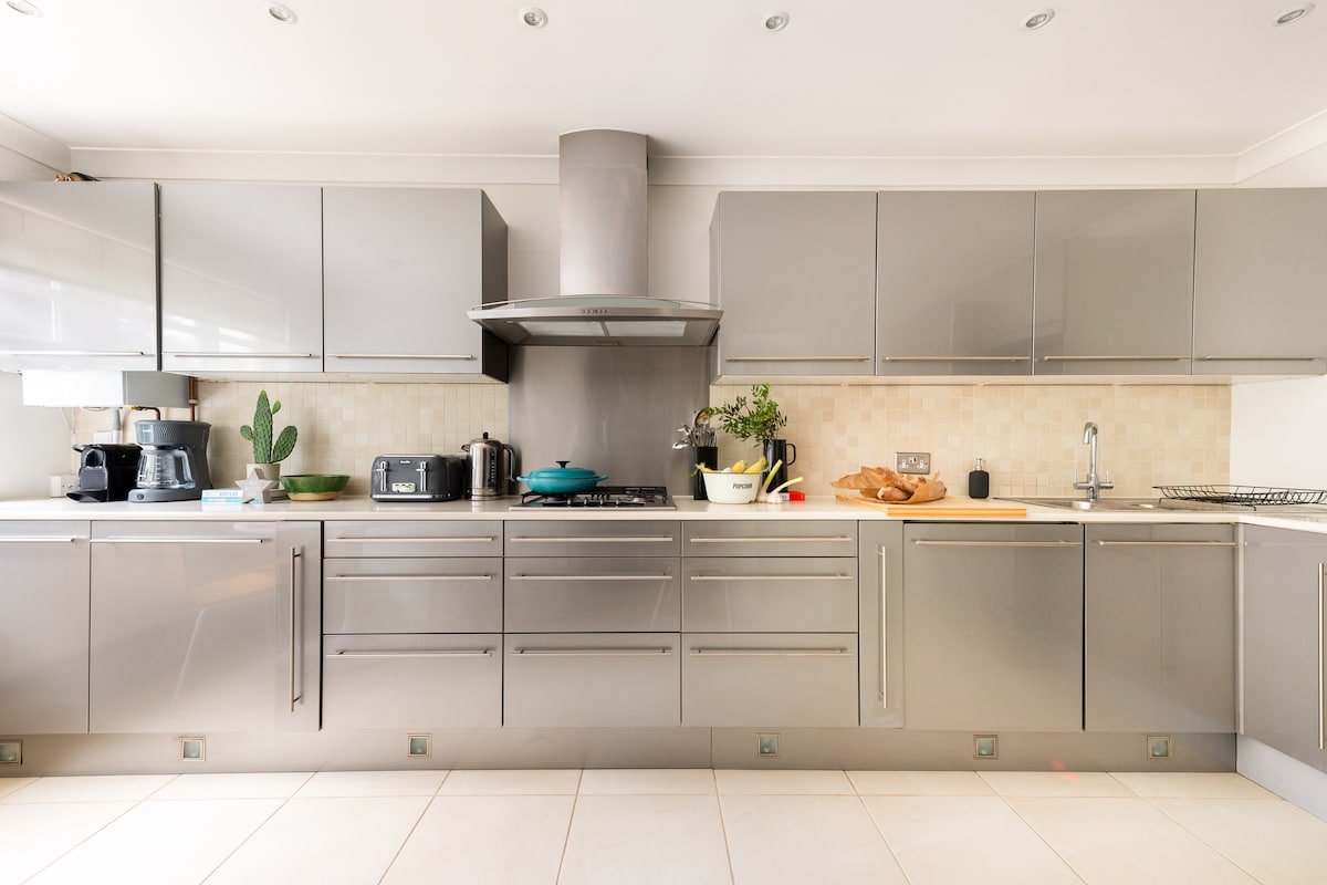 A modern kitchen is presented with sleek, glossy cabinets and ample countertop space. Various appliances, including a coffee maker and toaster, are arranged along the countertop. A decorative plant adds a touch of greenery, while the cooktop and hood are centrally located.