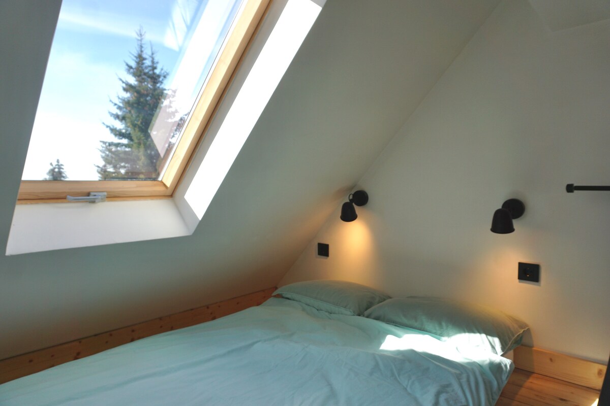 A cozy sleeping area features a sloped ceiling with a skylight allowing natural light to flood in. Soft bedding is arranged on a spacious bed, with stylish wall sconces providing gentle illumination on either side.