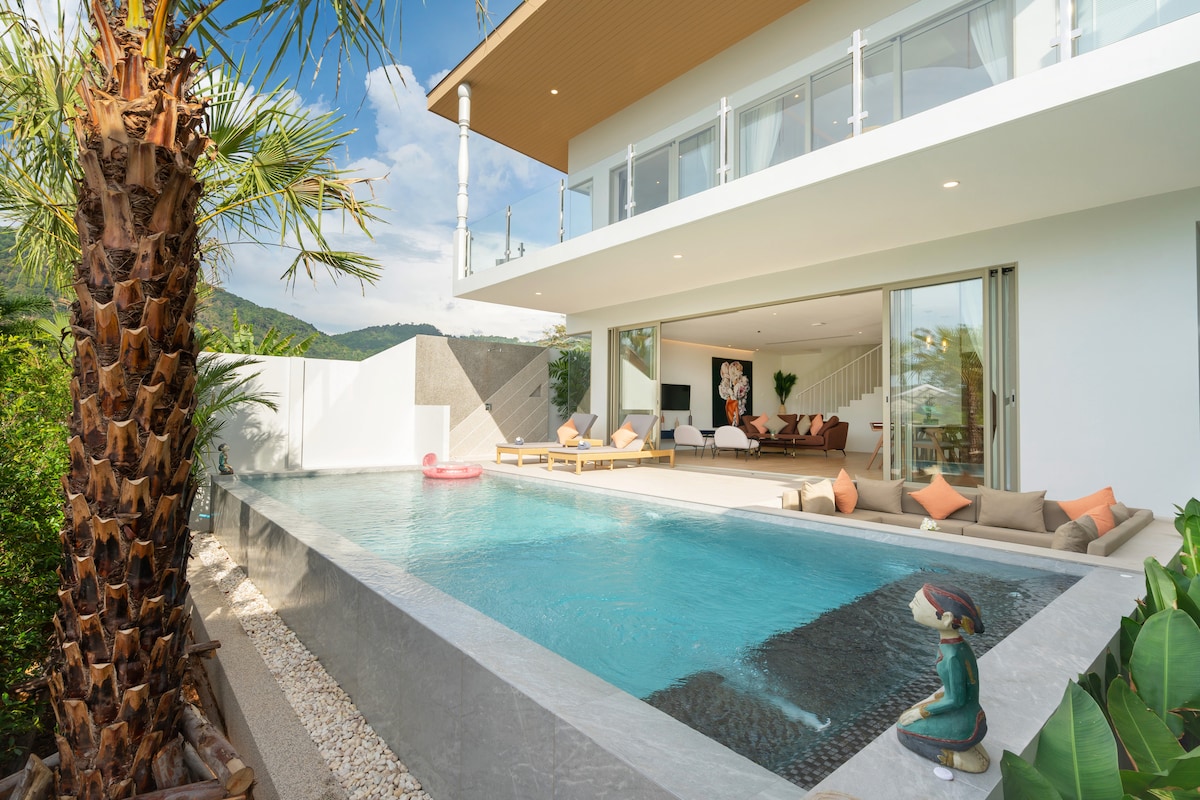 The villa showcases an inviting private pool, framed by lush greenery and a palm tree. A sunken seating area is nestled beside the pool, while large glass doors provide a view into the spacious living room. Natural light bathes the exterior, reflecting a serene atmosphere.