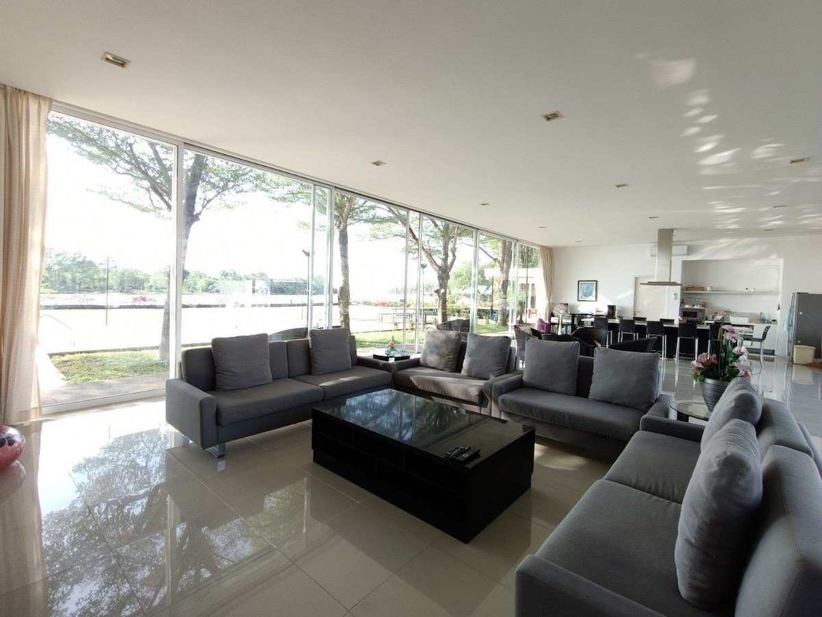 A spacious living area is filled with natural light from large glass windows. Comfortable grey sofas surround a low coffee table, creating an inviting gathering space. The open layout connects to a well-equipped kitchen, and outdoor views of greenery and the river enhance the atmosphere.