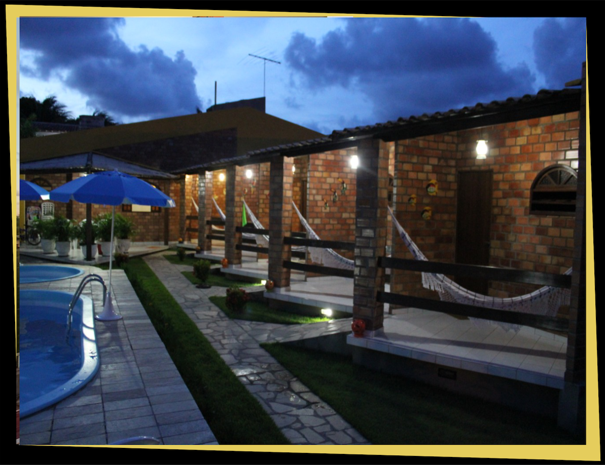 A serene evening scene captures a swimming pool surrounded by brick structures. Comfortable hammocks hang between wooden beams, while umbrellas provide shade over neatly arranged pathways. Soft lighting from wall fixtures enhances the inviting atmosphere of the outdoor space.