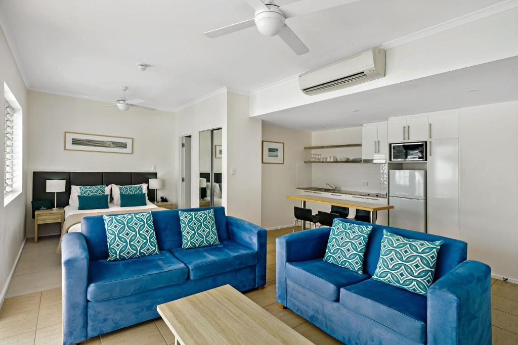 A bright and airy living space is featured, showcasing two blue sofas adorned with patterned cushions. A wooden coffee table sits in the center, while a kitchenette and dining area are visible in the background, along with a glimpse of a bedroom area beyond.