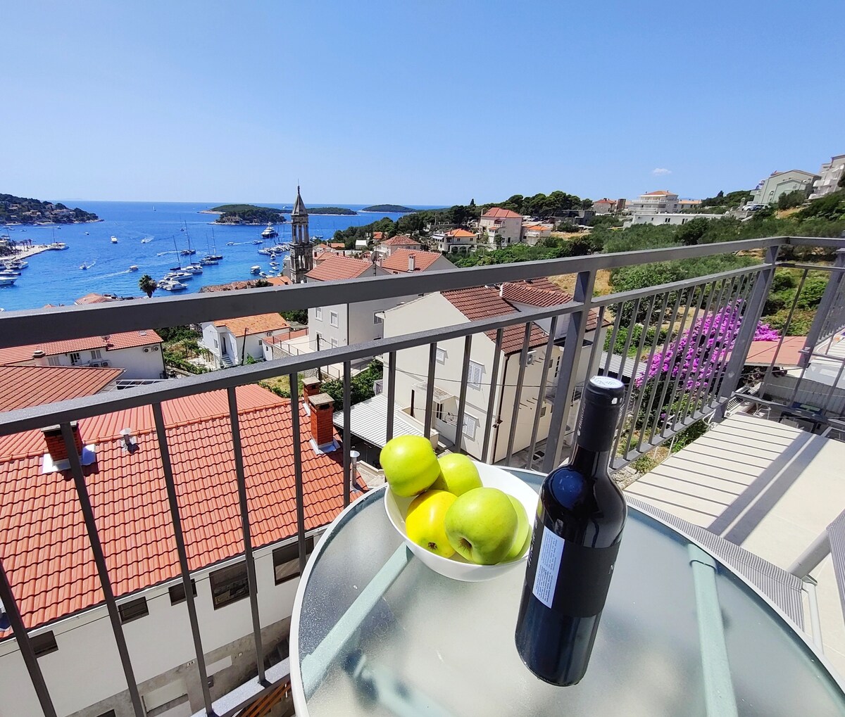 A private balcony is shown equipped with a glass table and two chairs. A bowl of green apples and a bottle of wine are placed on the table. The expansive view reveals the harbor, coastal landscape, and nearby rooftops under a clear blue sky.