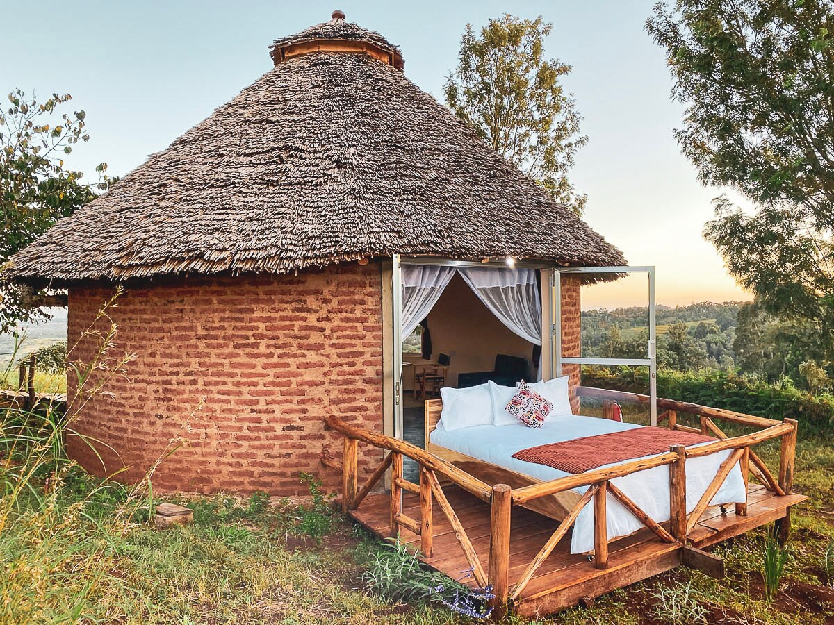 A circular eco-lodge features a thatched roof and wooden deck, surrounded by greenery. Open curtains reveal a spacious sleeping area with a large bed, while outdoor seating invites relaxation. Scenic views of the surrounding landscape are visible through large windows.