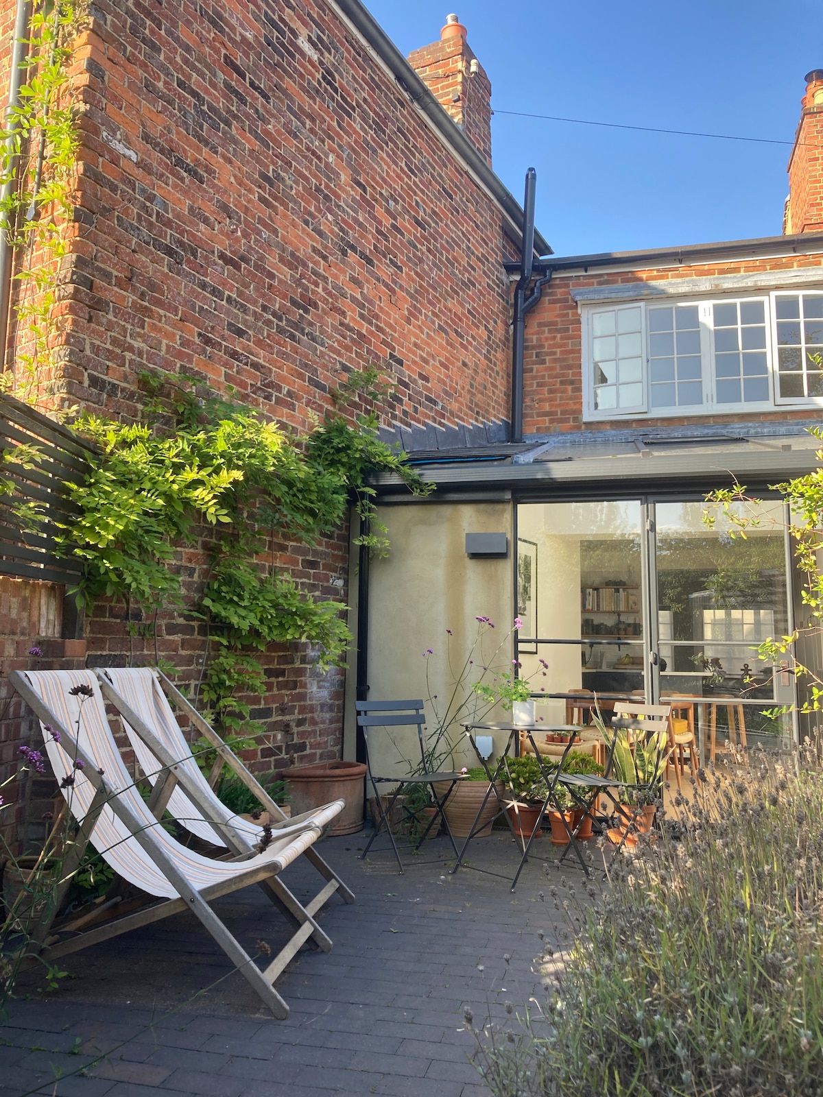 An outdoor patio area features comfortable loungers and a small dining set. Greenery climbs the brick wall, merging with the natural surroundings. Large glass doors provide access to the interior, allowing for an inviting connection between indoor and outdoor spaces.