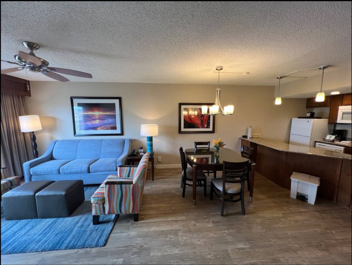 A living area showcases a light blue sofa and colorful accent chairs, complemented by a dining table with four chairs. A partial kitchen is visible, featuring white appliances and a stone countertop. Soft lighting and a patterned area rug enhance the space's comfort.