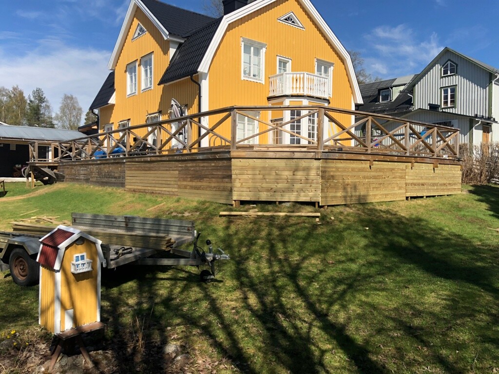 A charming yellow villa features a spacious wooden deck surrounded by a well-kept lawn. A small shed is positioned in the foreground, providing a cohesive look with the house. The property is enhanced by a clear blue sky casting light on the architectural details.