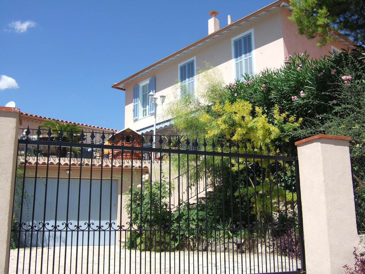 A charming pink house is seen behind a black wrought-iron gate, framed by lush greenery and colorful foliage. The second floor features light blue shutters and a balcony. The driveway leads to a garage, set against a clear blue sky.