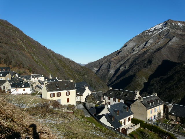 A view of a quaint village nestled in a valley, surrounded by rolling hills and mountains. The houses feature traditional architecture with sloping roofs. Clear blue skies contrast with the snow-capped peaks in the distance, highlighting the serene natural environment.