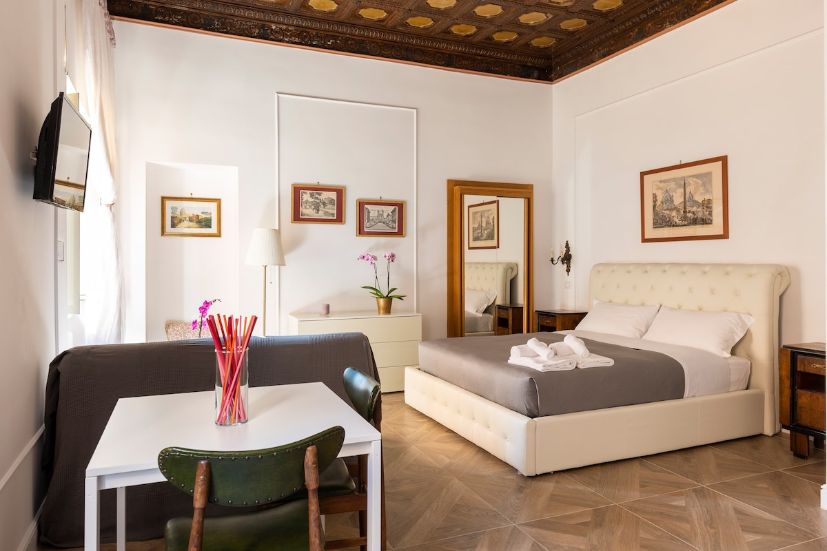 A bright and spacious room features a large bed with soft linens and a decorative headboard. A small dining table with green chairs complements the furnishings. Wall-mounted artworks and a tall lamp add character, while the ceiling showcases intricate wooden details.