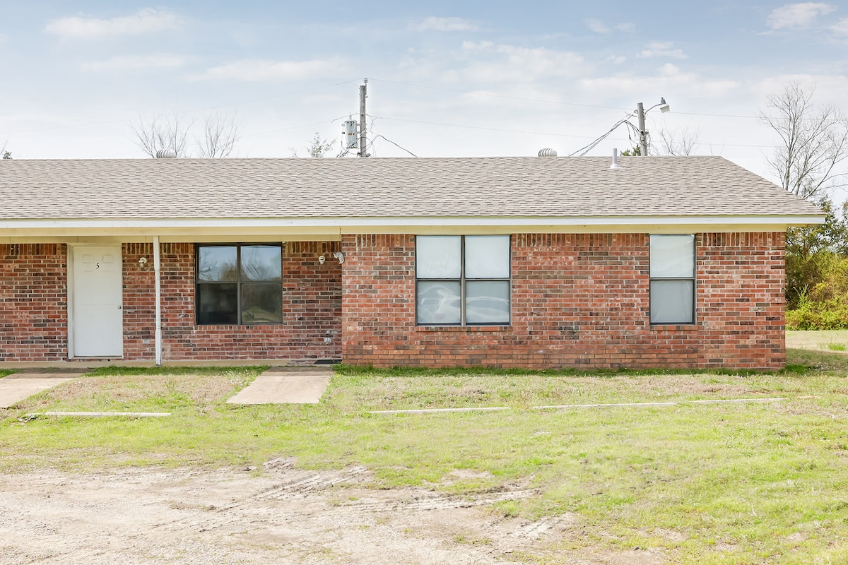 The exterior of a single-story brick building is shown, featuring a simple front entrance and two large windows that allow natural light inside. The well-maintained lawn and clear blue sky create an inviting look for the residence.