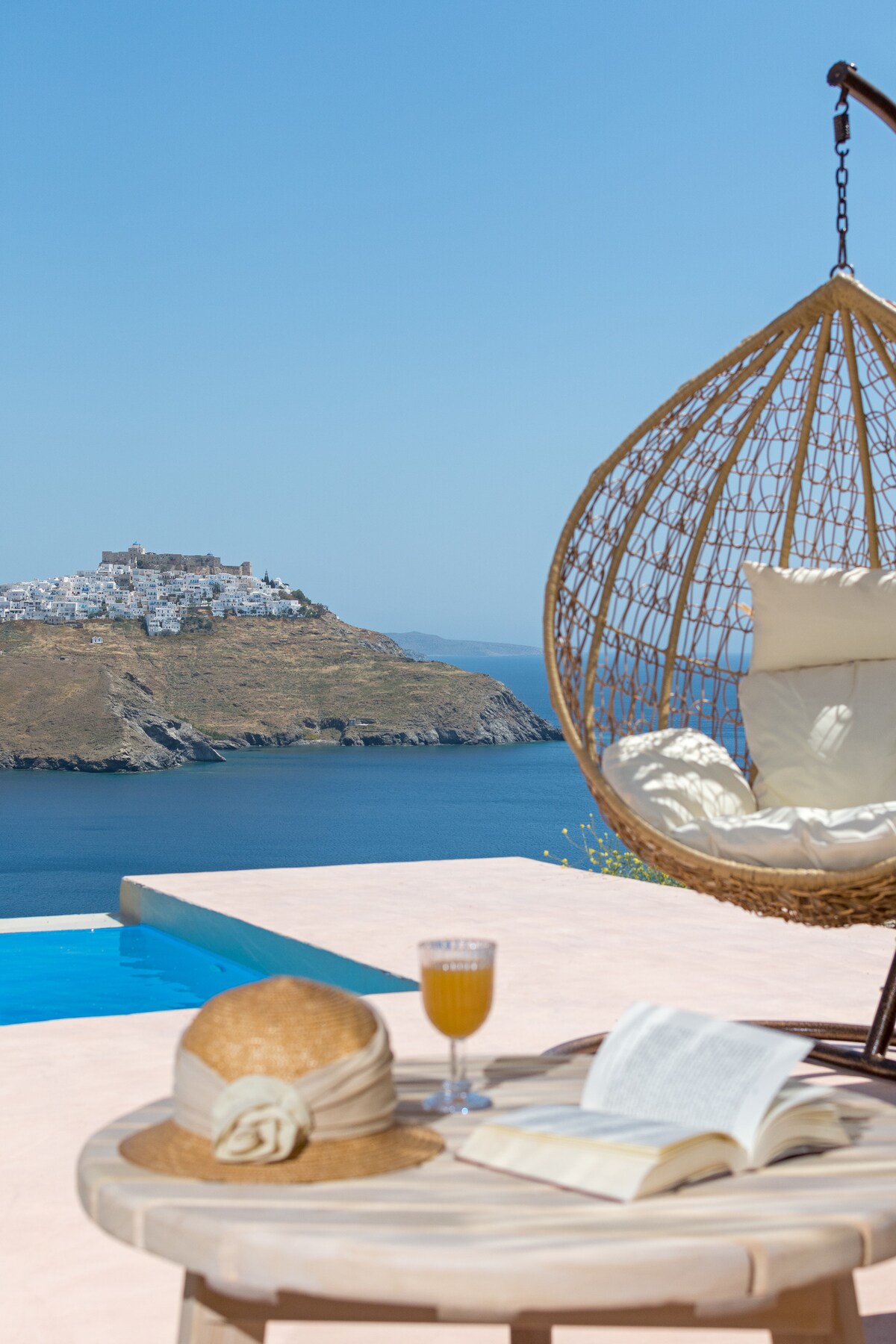A hanging chair is positioned by the pool, offering views of the sea and nearby island. A side table holds a book and a glass, while a straw hat rests on the table, enhancing the serene atmosphere.