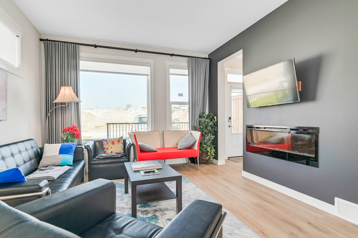 A modern living area features comfortable seating with a mix of dark and bright upholstery, complemented by large windows allowing natural light. A sleek wall-mounted TV is positioned above a stylish fireplace, enhancing the contemporary decor. Potted plants add a touch of greenery.