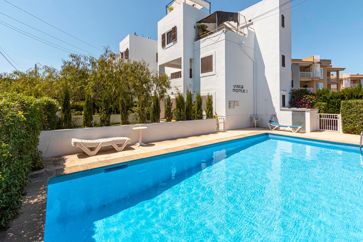A communal swimming pool is surrounded by lush greenery, with a sunlit deck featuring lounge chairs. The clear blue water reflects the bright sky, while a white building with several balconies stands in the background, harmonizing with the calm atmosphere of the area.