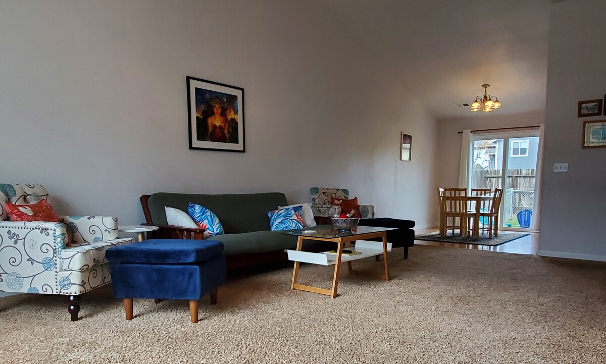 A comfortable living area features a variety of seating options, including a green couch and patterned armchairs. A wooden coffee table sits in the center, surrounded by plush ottomans. Light filters in through a doorway leading to an outdoor space, enhancing the welcoming ambiance.