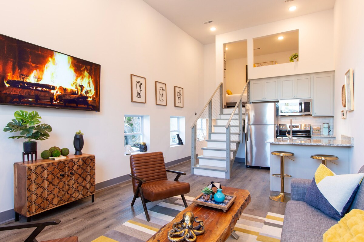 A chic living area features a warm fireplace display on the wall-mounted TV. A stylish wooden coffee table sits at the center, surrounded by comfortable seating. The modern kitchen and staircase can be seen in the background, enhancing the spacious design.