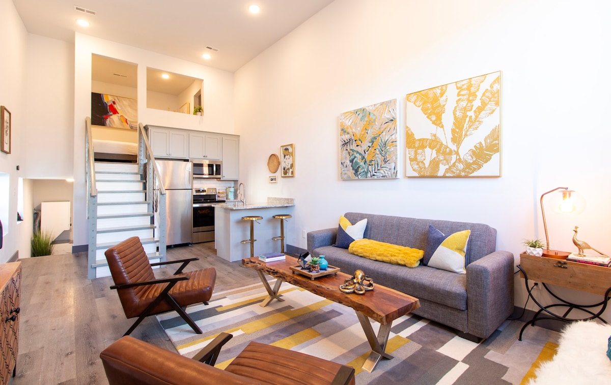 A spacious living area features a stylish gray sofa, accented by a yellow throw pillow. Two wooden chairs sit beside a coffee table, adorned with decorative items. The walls display framed art, and stairs lead to the lofted bedroom above, with an open kitchen visible in the background.