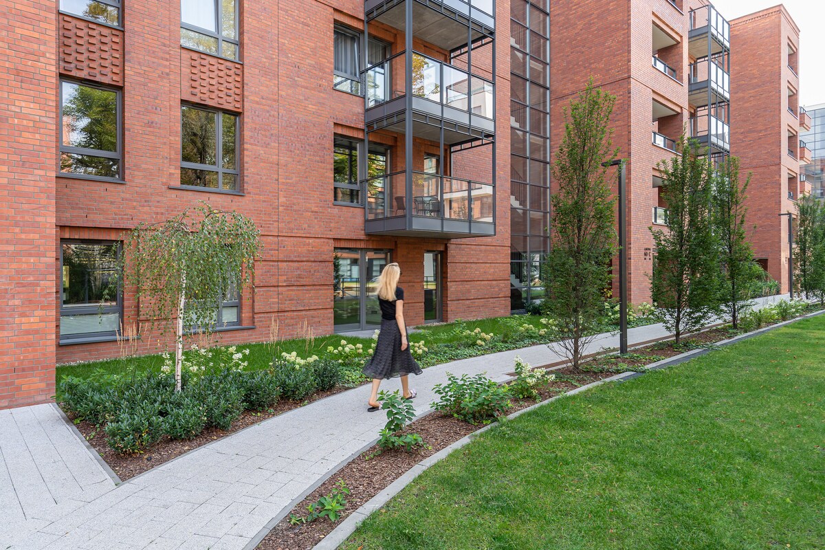 An elegant brick building with multiple balconies is surrounded by well-maintained green spaces. A pathway meanders through the garden, enhancing the inviting outdoor atmosphere. Lush greenery and flowering plants are visible, contributing to the aesthetic appeal of the area.