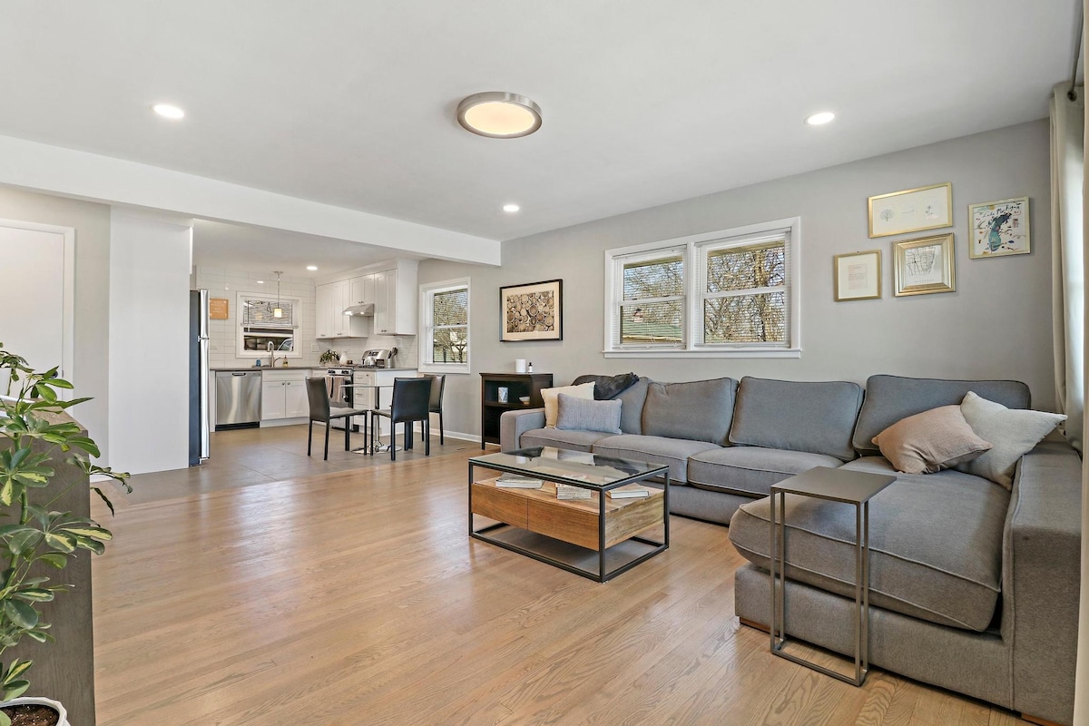 A light-filled living area is showcased, featuring a spacious sectional sofa and a modern coffee table with a wood accent. The open design leads to a dining space visible in the background, where a table with seating can be seen. Crisp white trim outlines multiple windows.