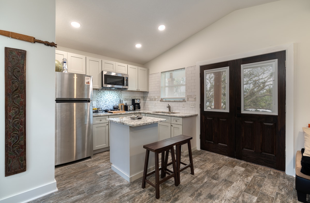 A modern kitchen is displayed, featuring stainless steel appliances and a central island with a wooden stool. Bright cabinetry complements the light countertops, while natural light streams through the large double doors leading outside.