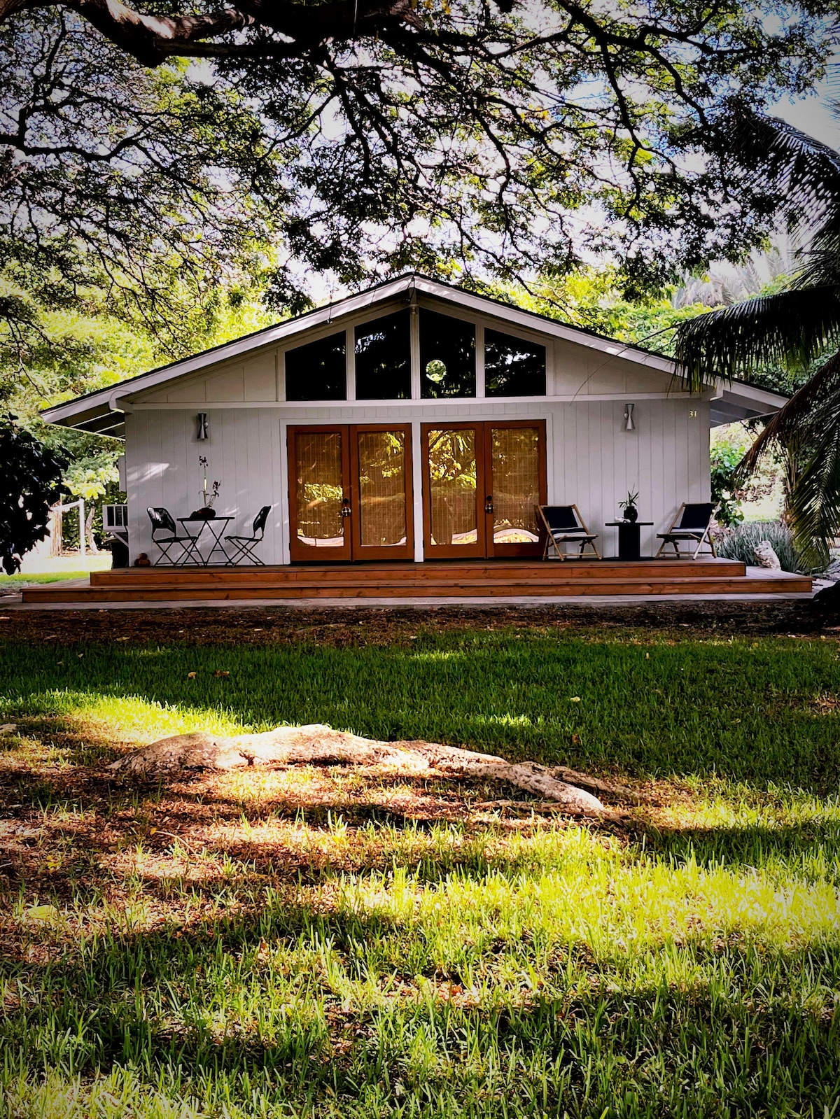 A midcentury modern cottage features large glass doors and a welcoming porch with two chairs and a small table. Surrounded by lush greenery, the structure is set against a backdrop of mature trees and a manicured lawn.