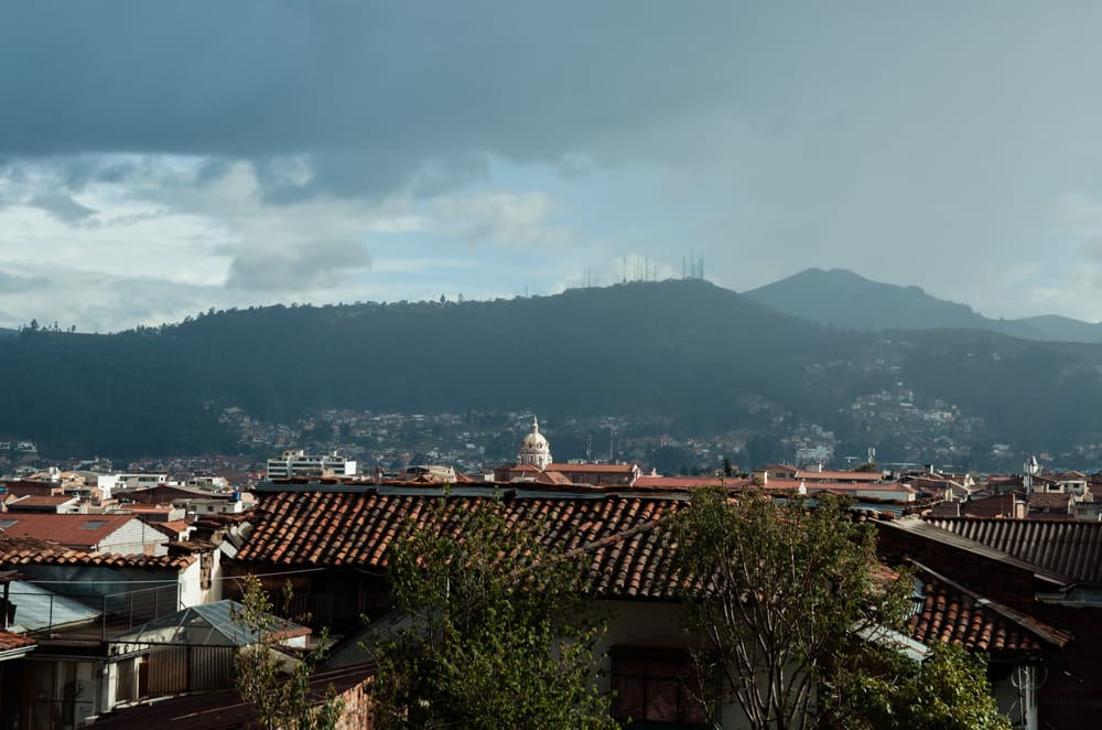 A panoramic view of the cityscape features red-tiled roofs complemented by lush greenery. The distant mountains are partially shrouded in clouds, while the prominent dome of a building is visible, highlighting the architectural richness of the area.