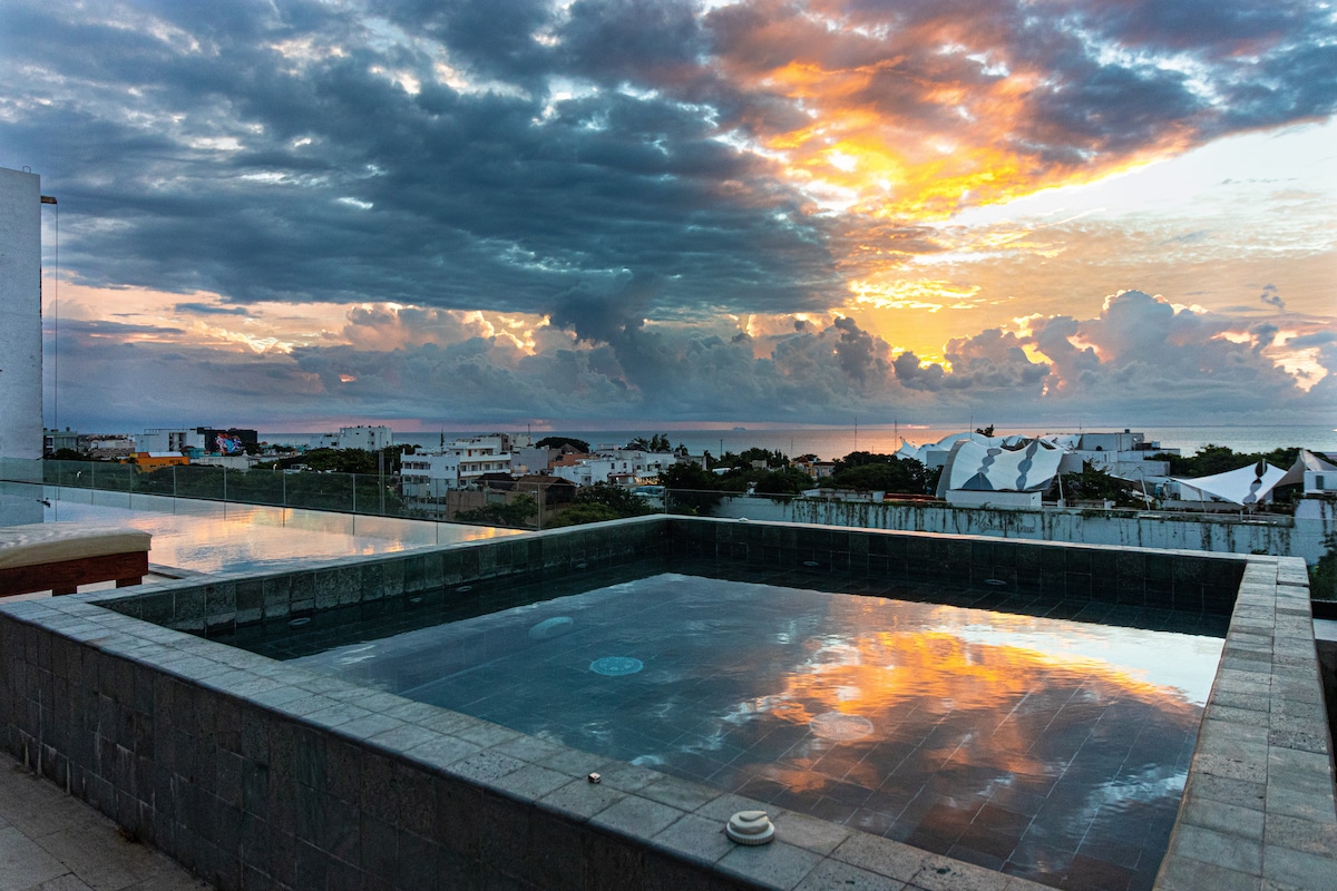 A rooftop area features a rectangular pool with dark stone tiles, reflecting a vibrant sunset. The sky shows clouds in hues of orange, pink, and gray. A view of the surrounding buildings and landscape is visible in the background.