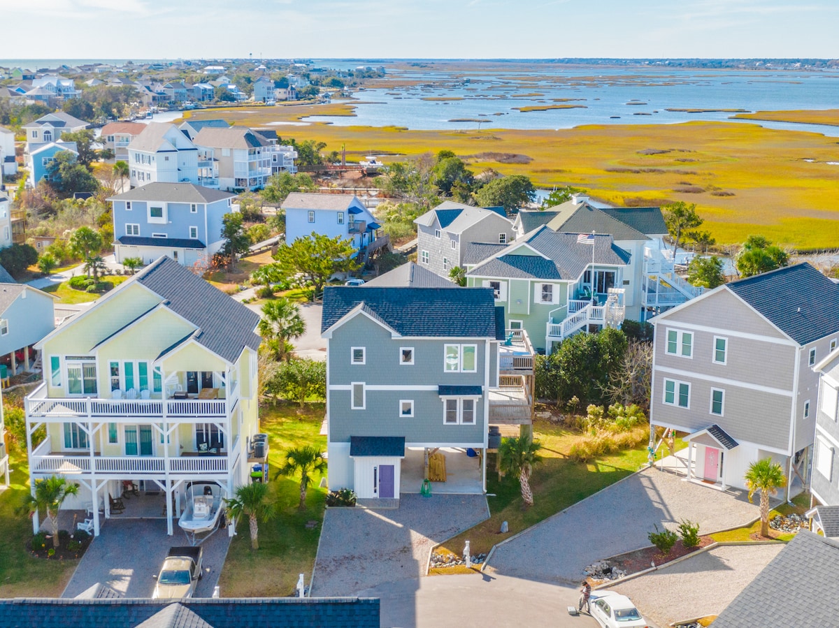 Aerial view of a tranquil coastal neighborhood showcases modern homes arranged around a winding street. Lush green lawns and sandy driveways are evident, while scenic water views reveal marshlands and blue skies, creating a peaceful atmosphere in the surrounding area.