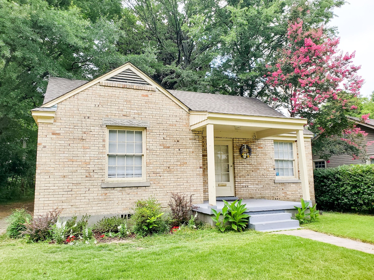 A charming single-story brick home is situated on a grassy plot, surrounded by trees and bright blooming flowers. The covered porch features a welcoming door with a seasonal wreath, while large windows allow natural light to filter into the living space.