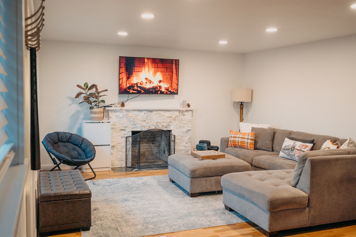A spacious living area features a comfortable sectional sofa and an accent chair. A flat-screen television displays a warm fireplace scene. Soft lighting highlights the stone fireplace, while a patterned rug adds texture to the hardwood floors.