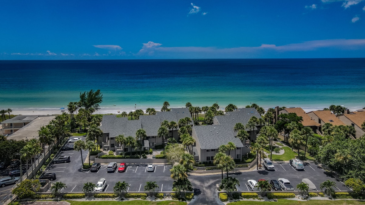 An aerial view captures the beachfront property bordered by lush palm trees. The ocean's blue waves gently meet the sandy shore, creating a serene backdrop. The well-maintained parking area accommodates several vehicles, highlighting the community's proximity to the beach.