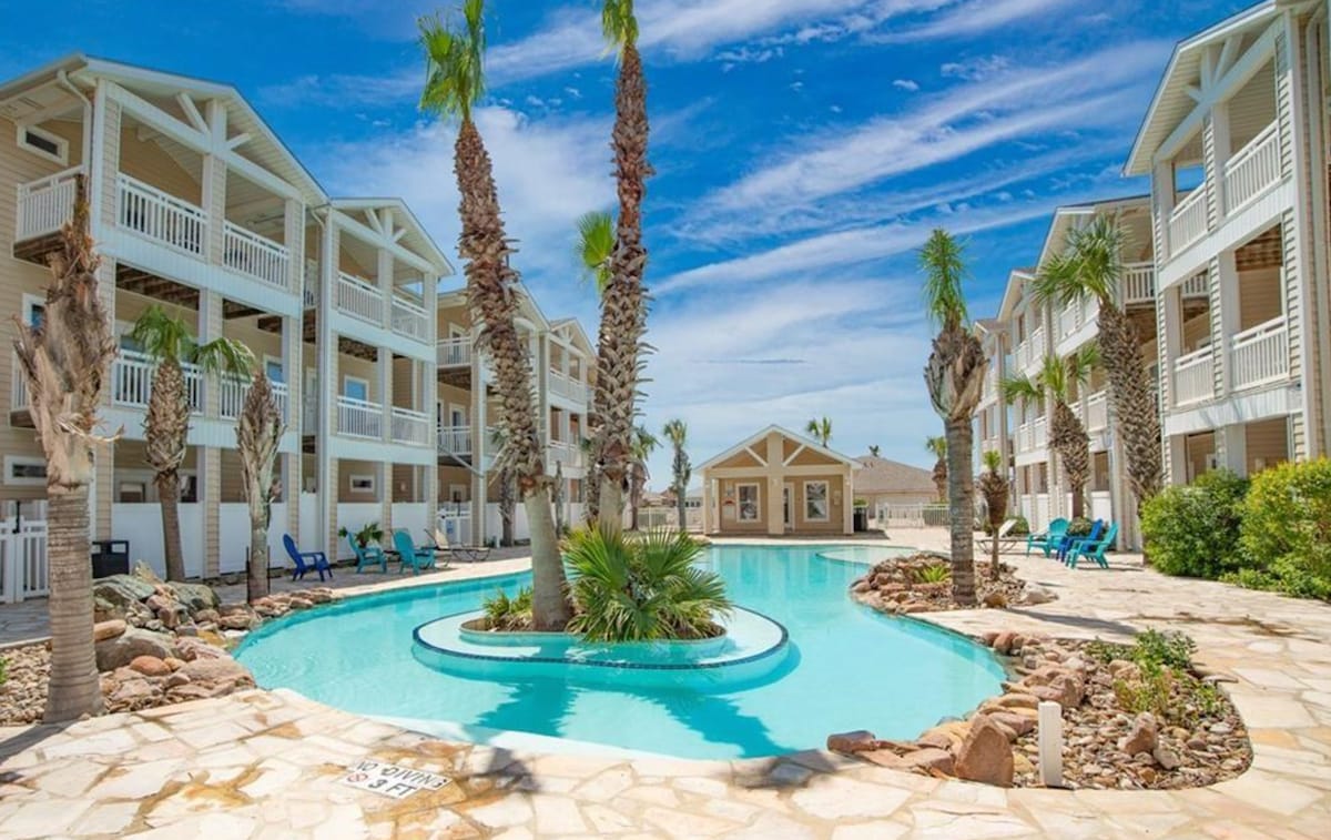 A tranquil outdoor pool is surrounded by palm trees and landscaped areas, creating a relaxing atmosphere. Nearby, two three-story buildings provide access to the pool area, with blue lounge chairs arranged on the shaded stone deck under a clear sky.
