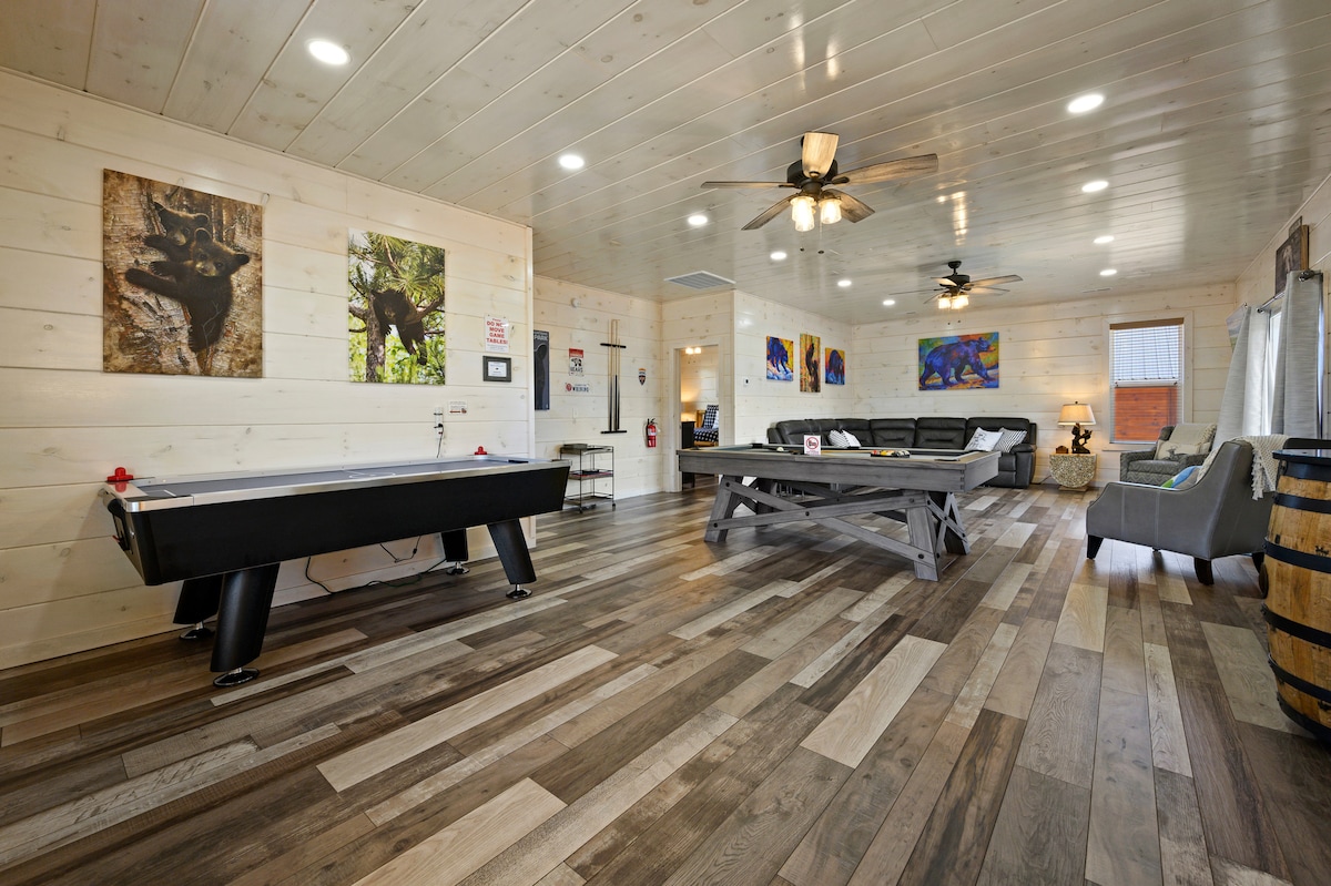 The spacious game room features a pool table and an air hockey table, with wooden flooring adding warmth to the space. Colorful artwork decorates the light wood walls, and large windows provide natural light. Comfortable seating areas are arranged for relaxation and enjoyment.