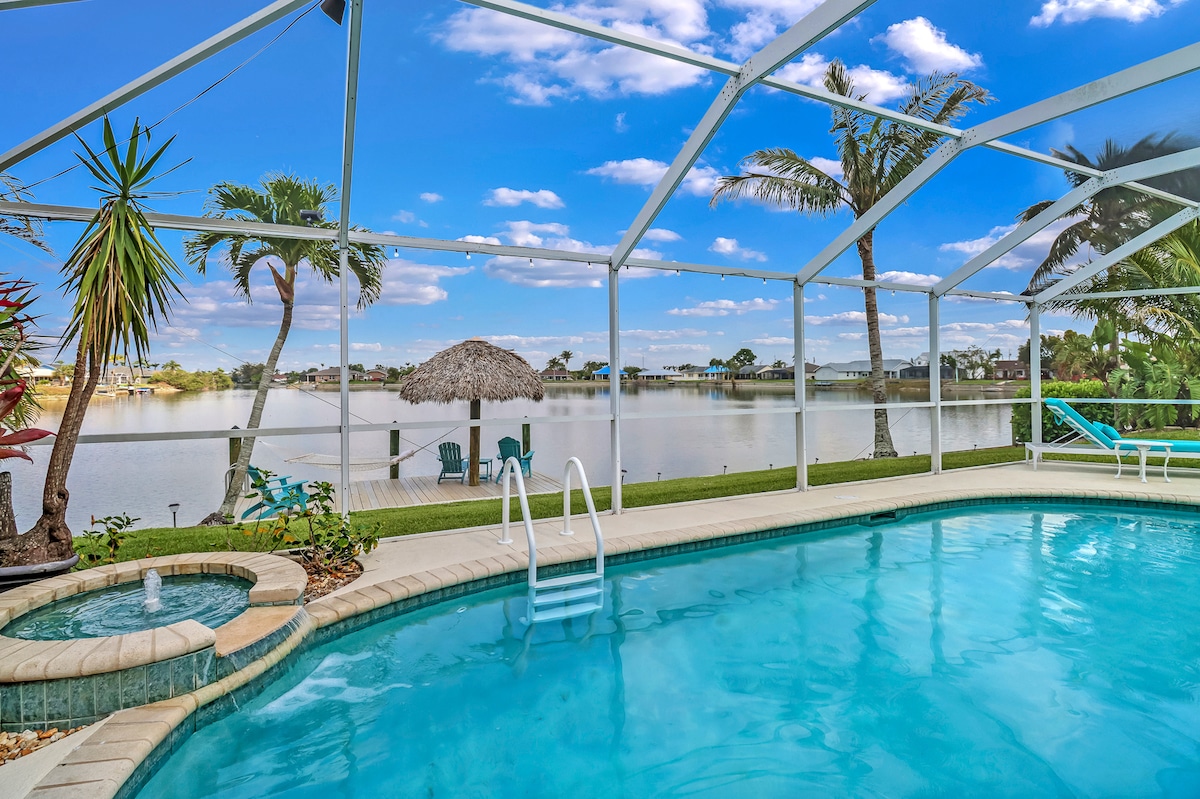 A tranquil pool area is highlighted by a clear blue swimming pool, with steps leading in. Lounge chairs are positioned nearby, and a shaded palapa provides a seating area, all overlooking a serene lake view framed by tropical plants and palm trees.