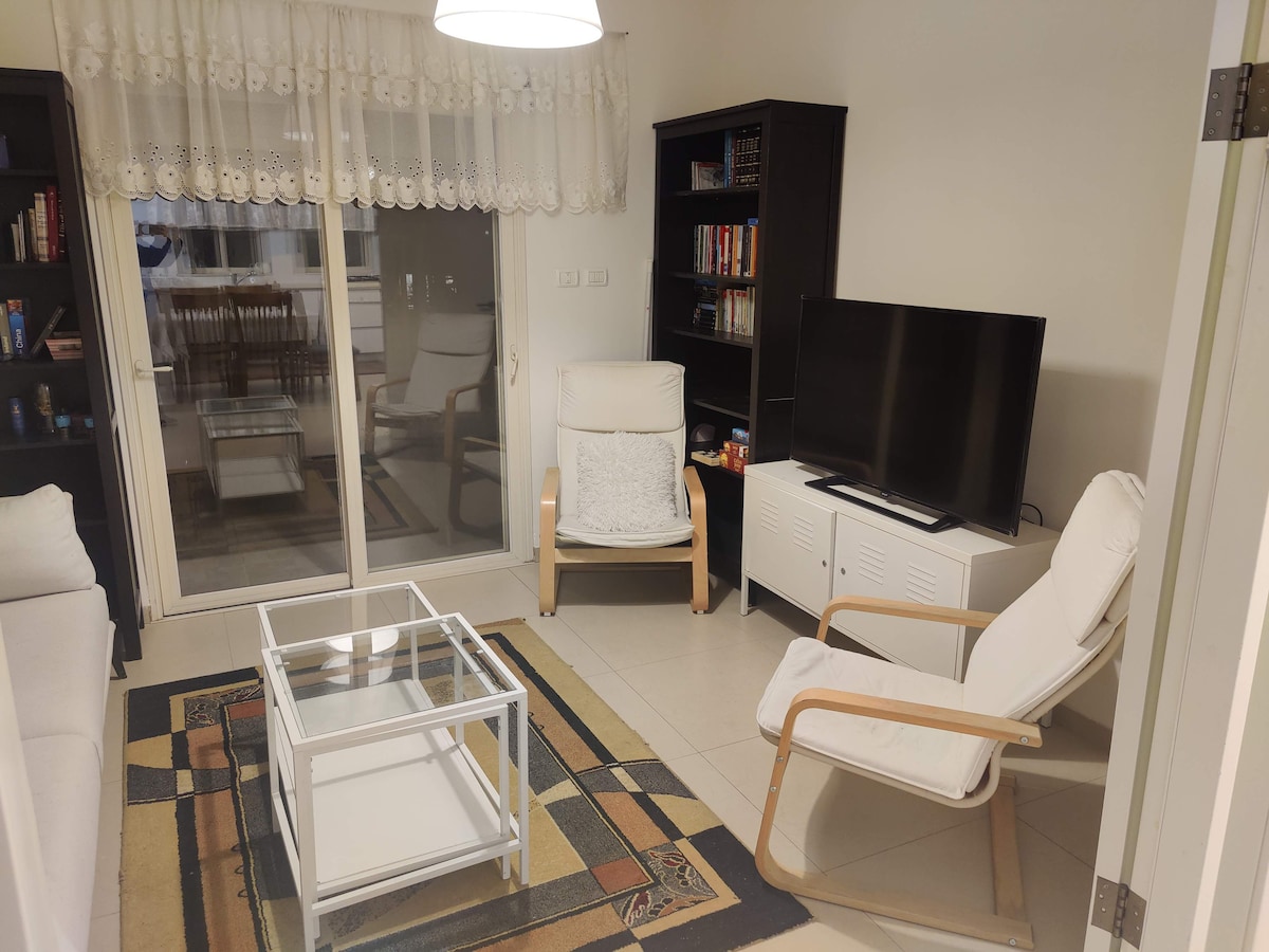 A cozy living area is presented with a glass coffee table and two white lounge chairs. A television sits atop a sleek media console, while shelves hold various books. Natural light enters through large sliding doors, offering a view of the outdoor space.