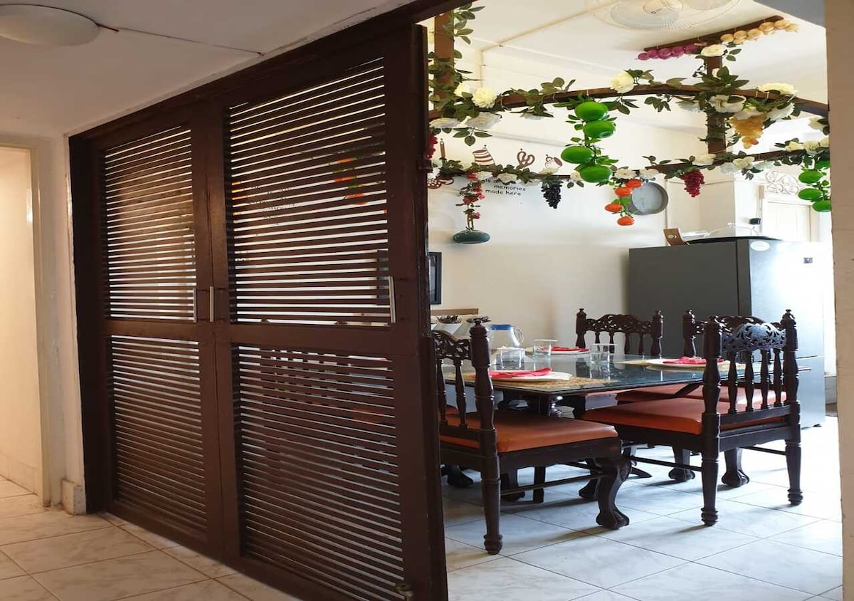 A dining area is viewed through wooden slatted doors, featuring a large table surrounded by six ornate chairs. The room includes a refrigerator and is decorated with hanging artificial greenery and fruits, adding a touch of color to the space.