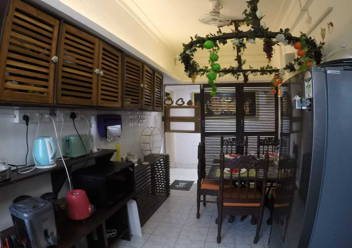 The kitchen area features a combination of wooden cabinets and a dining space with several chairs arranged around a table. Decorative elements include a ceiling piece adorned with greenery and colorful fruit accents. Appliances like a microwave, kettle, and refrigerator are visible.