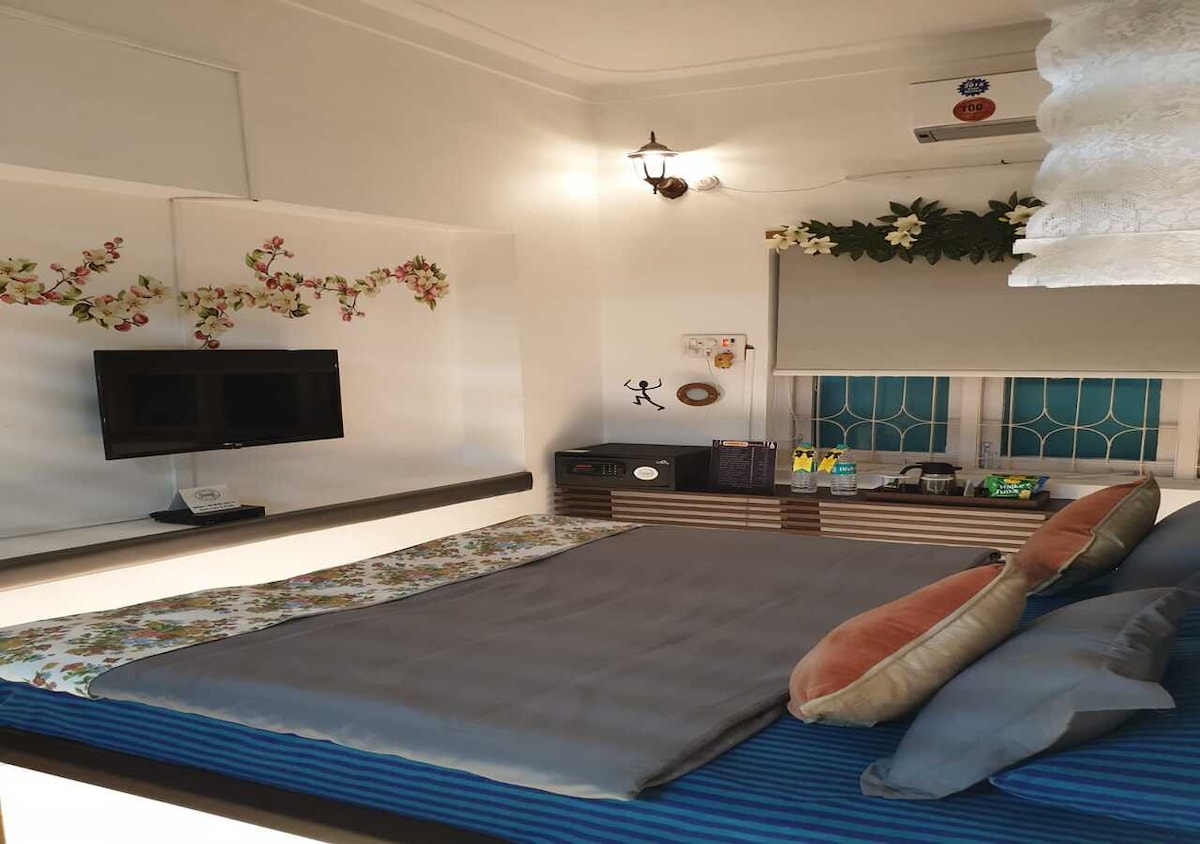 A comfortable room features a spacious bed covered with a blue striped blanket and decorative pillows. A flat-screen television is mounted on the wall, and a small counter displays a selection of snacks and drinks. Natural light filters through a partially covered window adorned with floral accents.