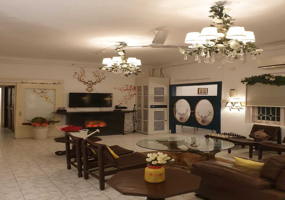 A spacious lounge area features a combination of wooden chairs and a leather sofa, arranged around a glass coffee table. Decorative lights, including a chandelier and floral accents, adorn the ceiling. A TV is mounted on the wall, and a faux fireplace adds a cozy touch.