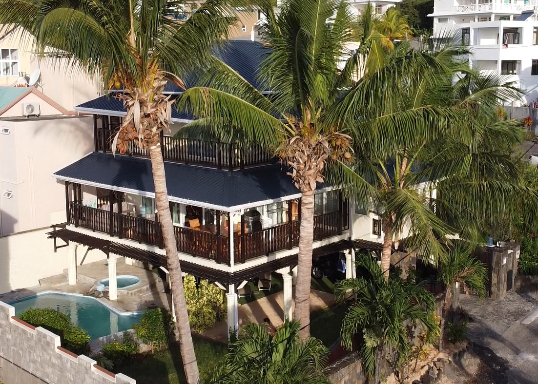 A two-story villa is surrounded by palm trees, with a private pool visible in the front. The open-air terrace showcases seating and dining areas. Large windows reflect the sun, offering a glimpse of the inviting interior. Lush greenery enhances the serene atmosphere of the property.