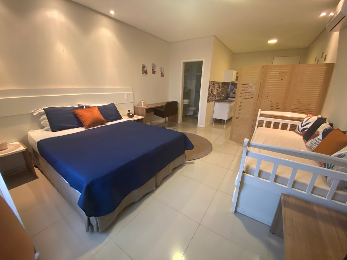 A well-arranged sleeping area features a spacious bed with a navy blue cover and contrasting orange accents. Natural light fills the room, highlighting a cozy seating area with a light-colored daybed and modern furnishings. A decorative partition offers privacy while maintaining an open feel.